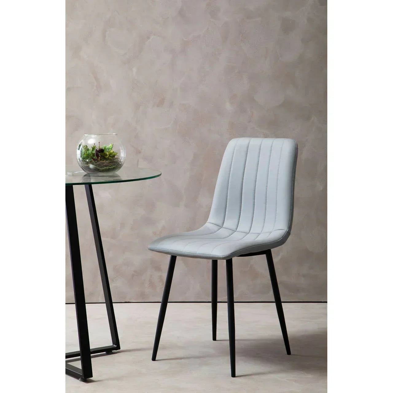 Grey dining chair with vertical stitching and black metal legs beside a round glass table