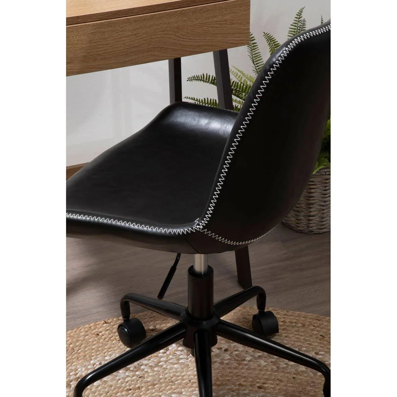 Black faux leather office chair with white stitching on castor wheels beside wooden desk