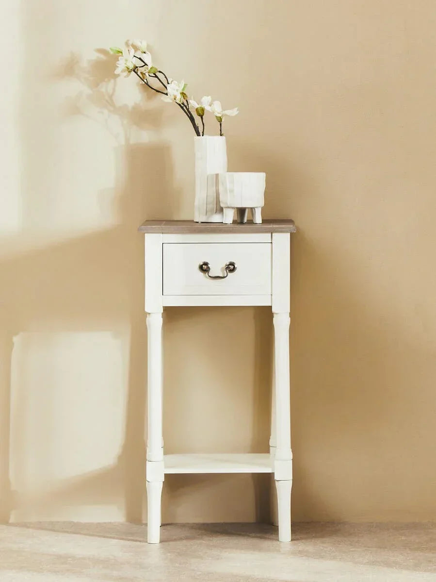 White wooden bedside table with a drawer and vases, against a neutral wall