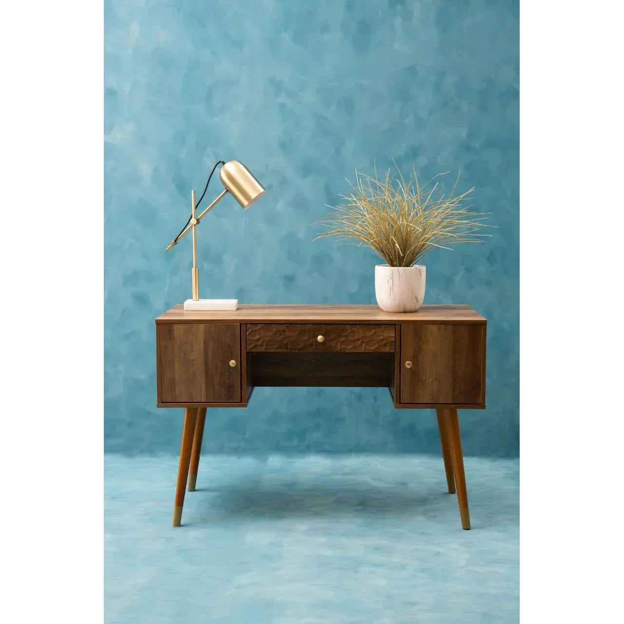 Mid-century wooden desk with gold lamp and potted plant against blue background