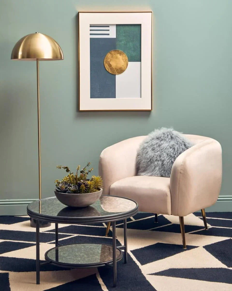 Modern beige armchair with gold legs beside round glass coffee table and brass floor lamp
