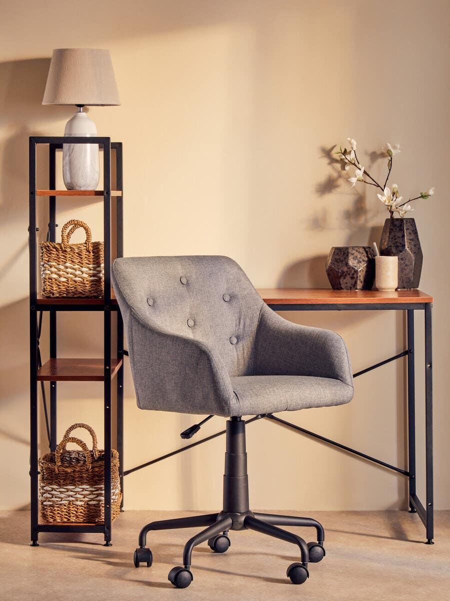 Grey office chair with castors at wooden desk beside black shelving unit and table lamp