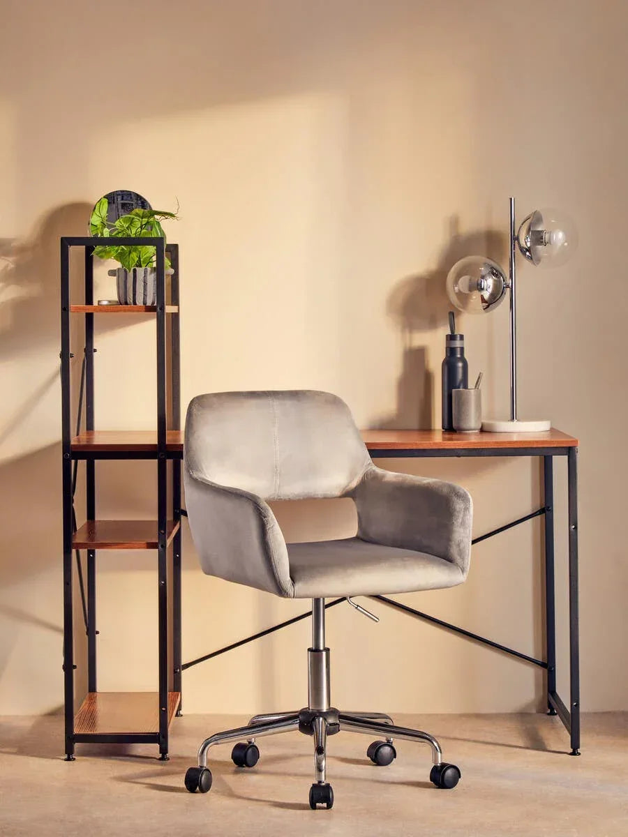 Grey velvet office chair with wheels in front of wood desk and black metal shelf unit