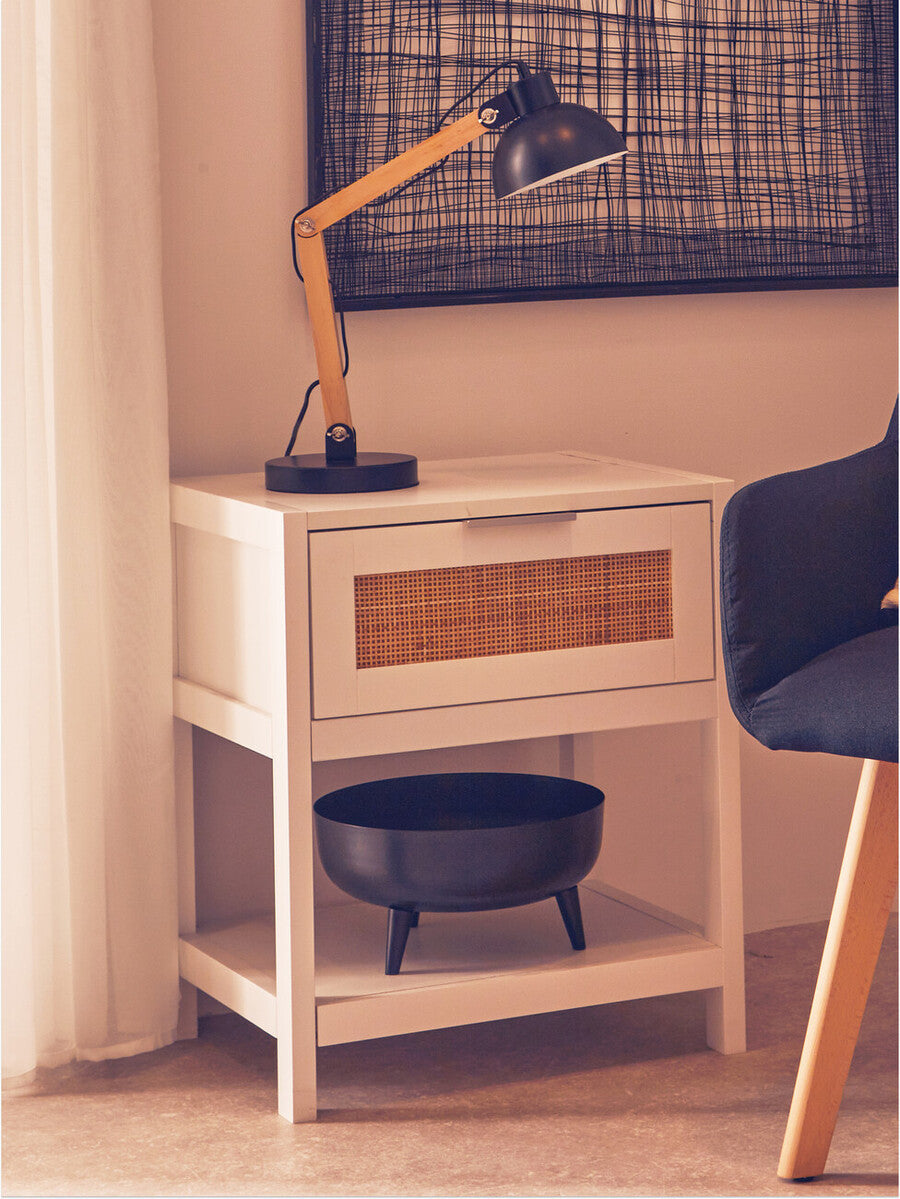 White wood side table featuring a rattan drawer front, a single drawer, and a lower shelf. A black lamp with a wooden arm is positioned on top, alongside a round black bowl. The table is set against a light-colored wall with a patterned artwork above it and a dark chair nearby. Natural light filters through sheer curtains