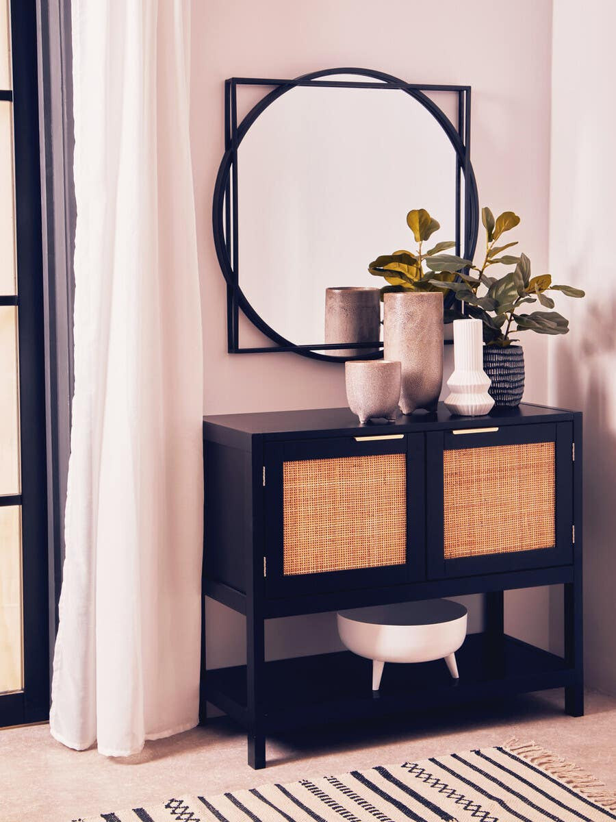 Black wood and rattan two-door side cabinet with a sleek design, featuring two rattan-woven doors and an open shelf below. A round mirror hangs above the cabinet, accompanied by decorative vases and greenery. The cabinet is positioned against a light-colored wall, with a patterned rug underneath