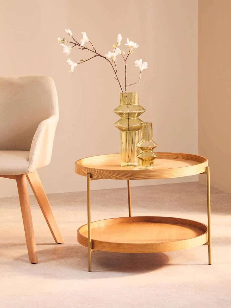 Round wooden coffee table with lower shelf and gold legs beside beige accent chair