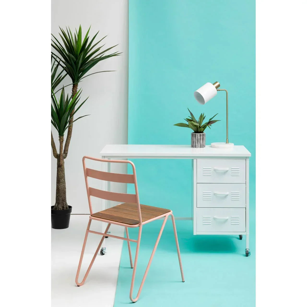 White metal desk with drawers, pink chair, table lamp and plant against blue background