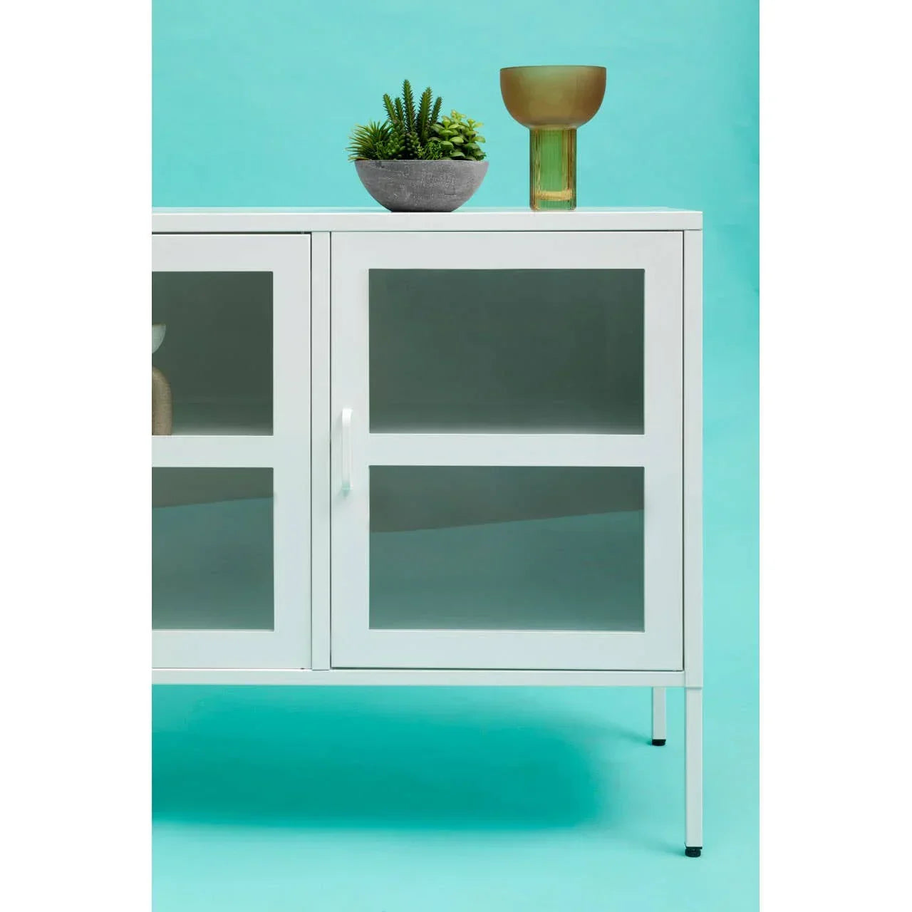 White storage cabinet with glass doors, potted plant, and green glass vase on blue background