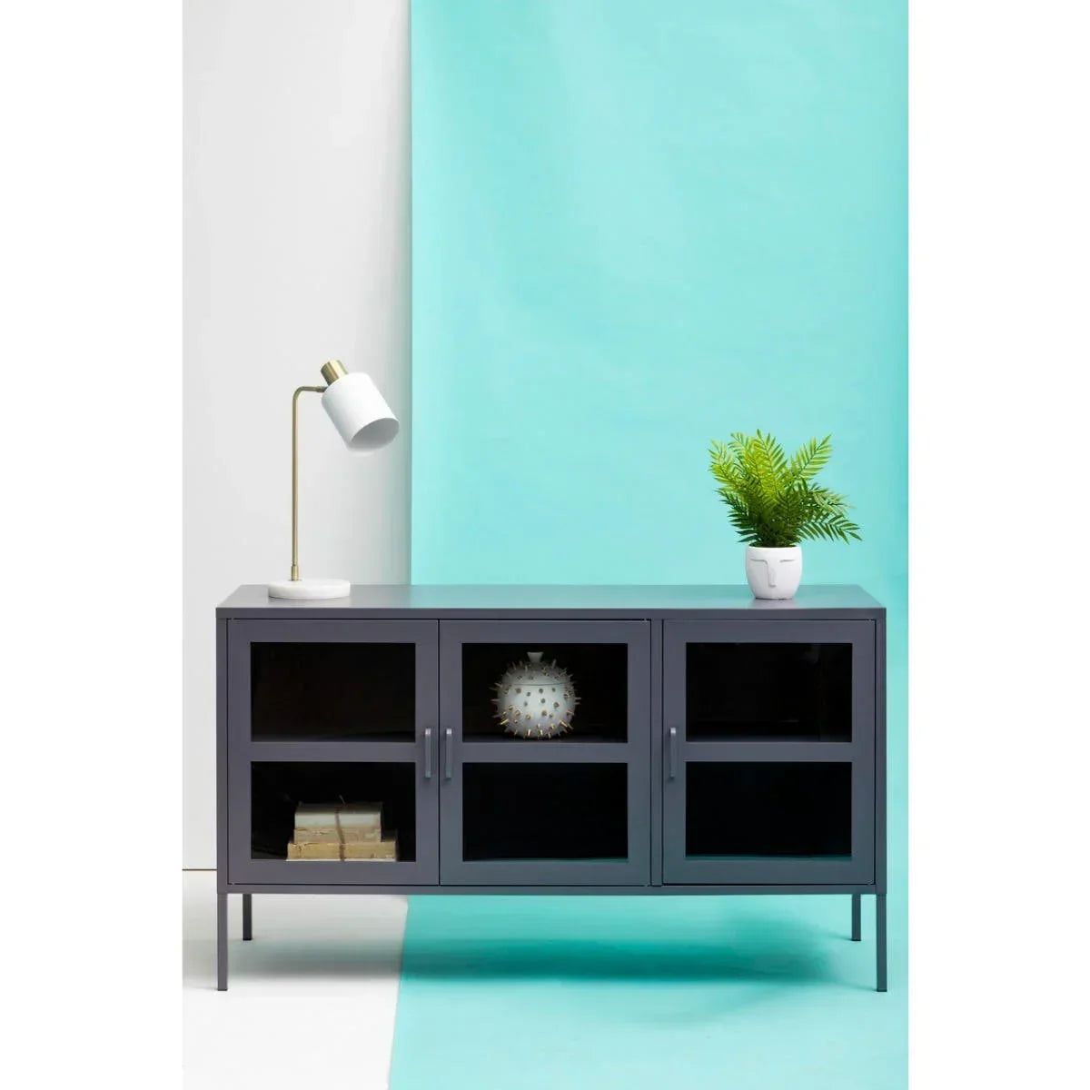 Grey metal sideboard with glass doors, table lamp, and potted plant against blue backdrop