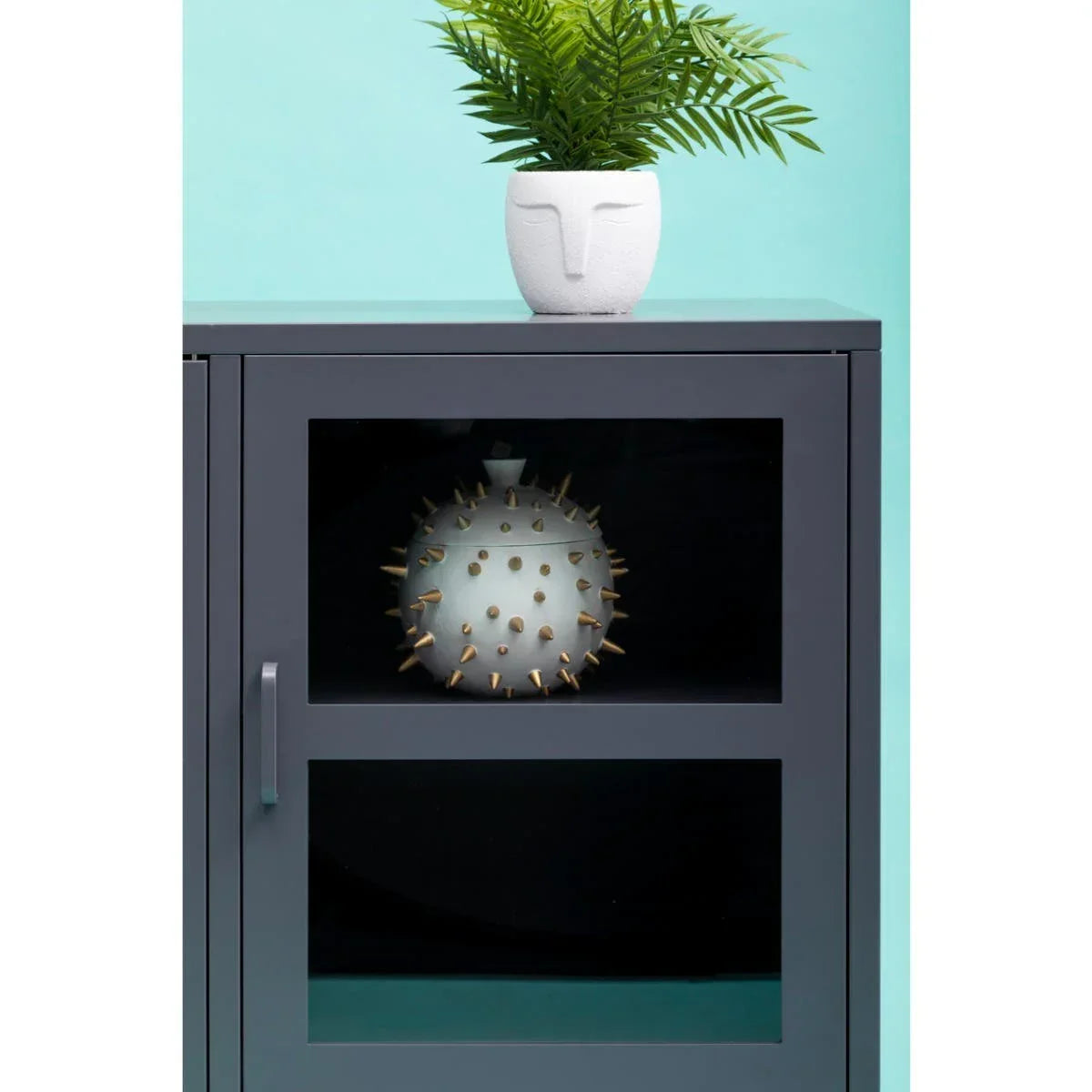 Grey cabinet with glass door, face planter with fern on top, spiked ceramic vase inside