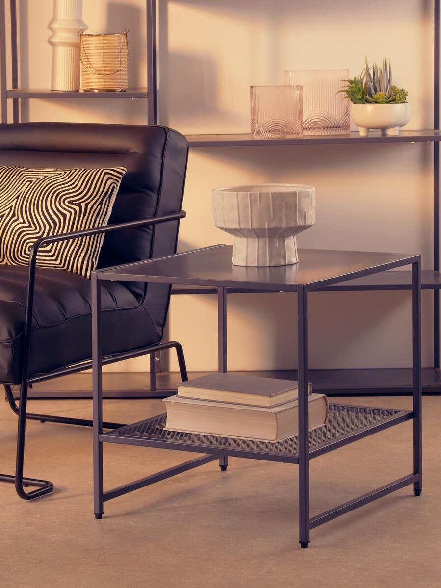 Black metal side table with lower mesh shelf beside leather armchair and decorative bowl