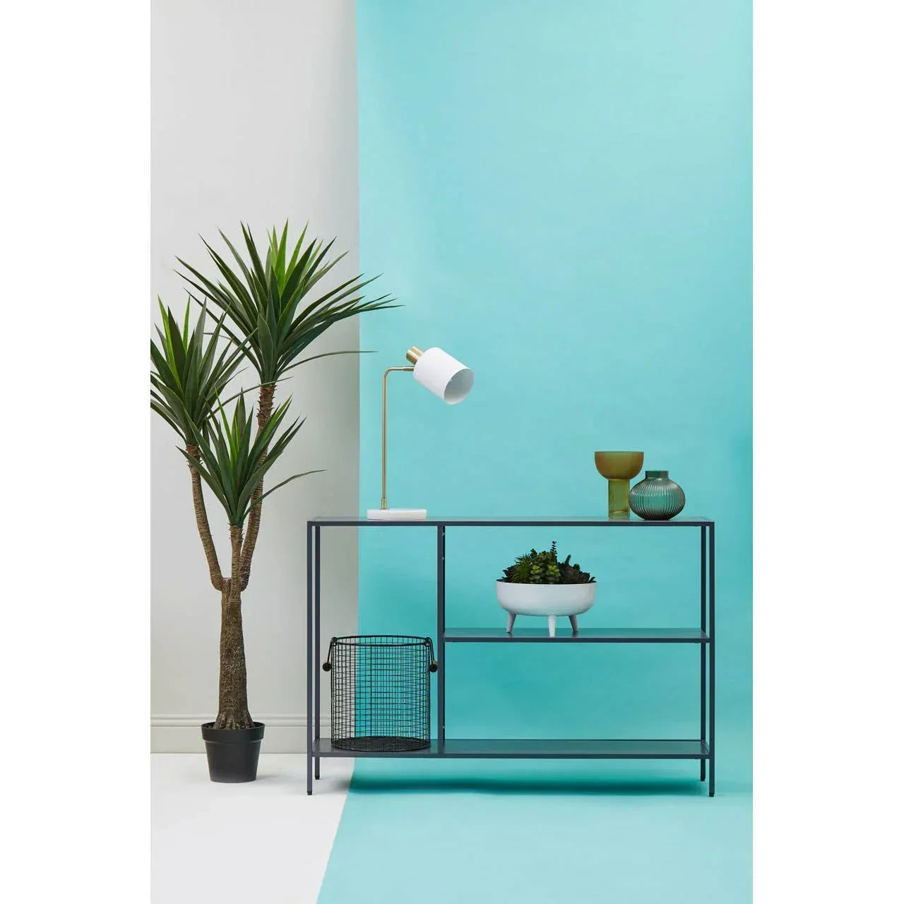 Black metal shelving unit with houseplant, lamp, glass vases, and wire basket on blue backdrop