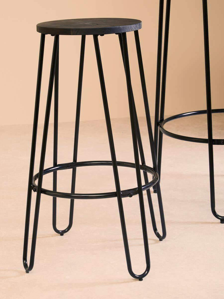 A black metal bar stool with a round seat and slender legs, featuring a minimalist design. The stool is positioned next to a tall bar table, showcasing the set's modern aesthetic against a neutral background