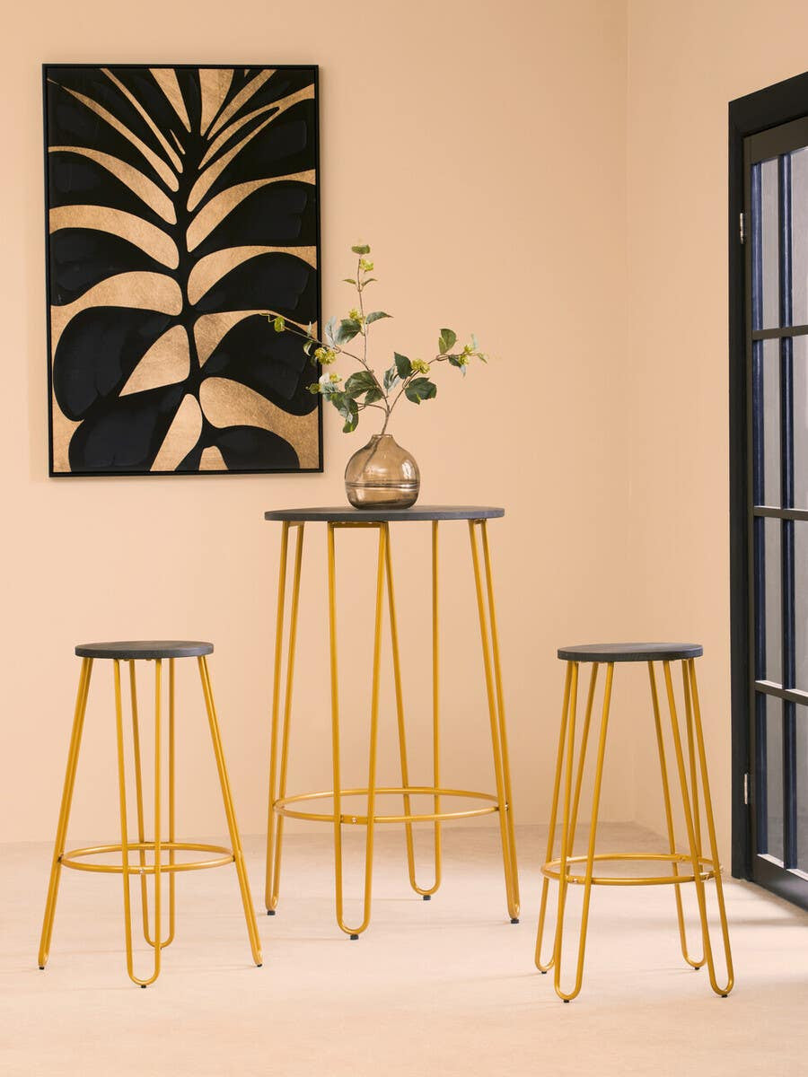 A three-piece bar table and stool set featuring a gold finish with sleek, modern design. The set includes one tall bar table and two matching stools, all with black tops. The background showcases a minimalist wall art piece with a plant design and a light-colored wall, creating a stylish and contemporary atmosphere