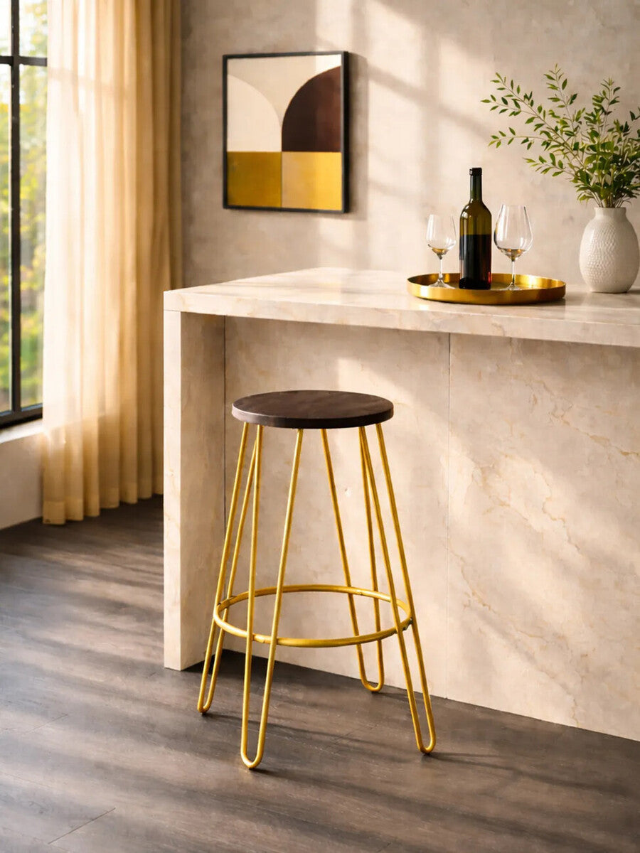 Gold finish metal frame bar stool with a round brown seat, positioned next to a light-colored countertop. A bottle of wine and a small tray are on the countertop, with a decorative mirror and a vase of flowers in the background. Natural light filters through a window, casting soft shadows on the wooden floor
