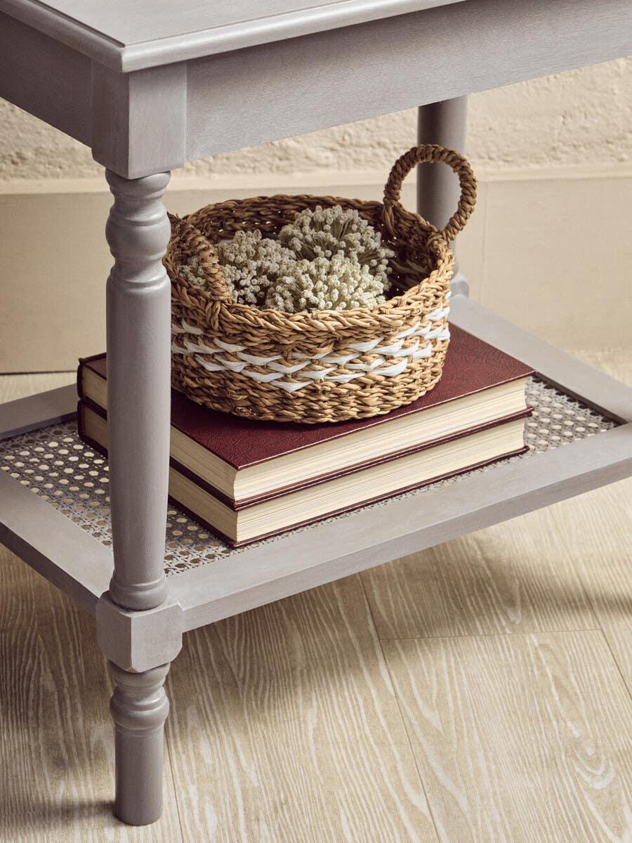 Grey wooden side table with lower shelf holding wicker basket and books on wood floor