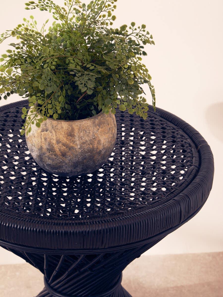 Black rattan side table with woven top and decorative plant in a stone pot