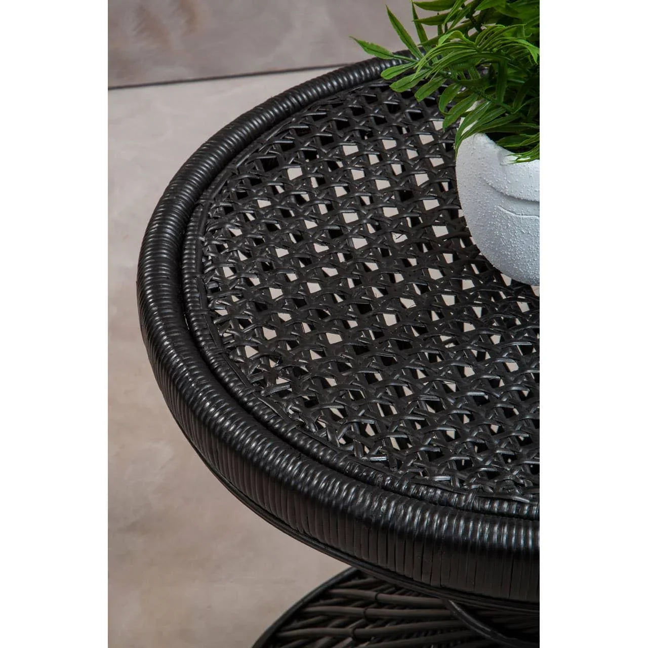 Black rattan side table with woven top and potted plant on light stone floor
