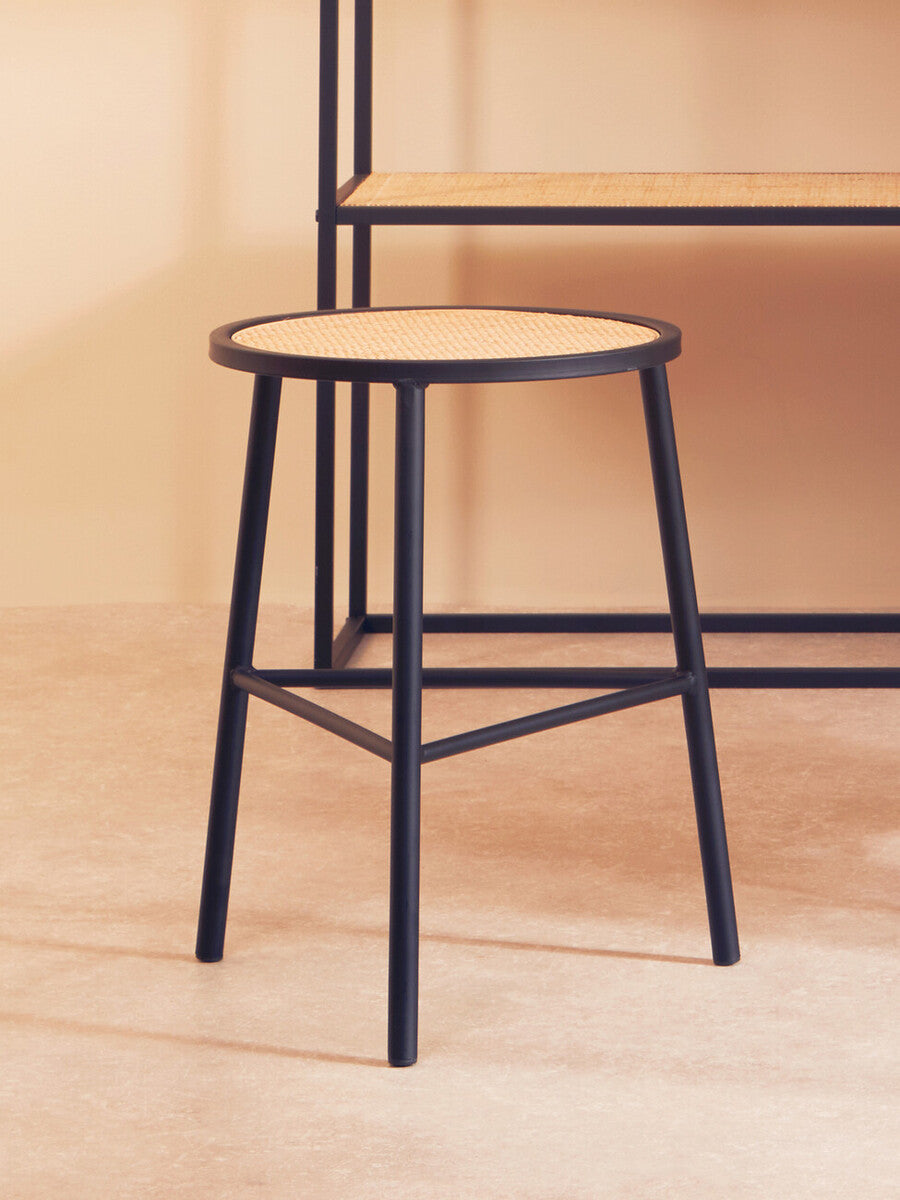 Rattan seat stool with a round top and black metal legs, positioned on a light wooden floor, with a minimalist shelving unit in the background