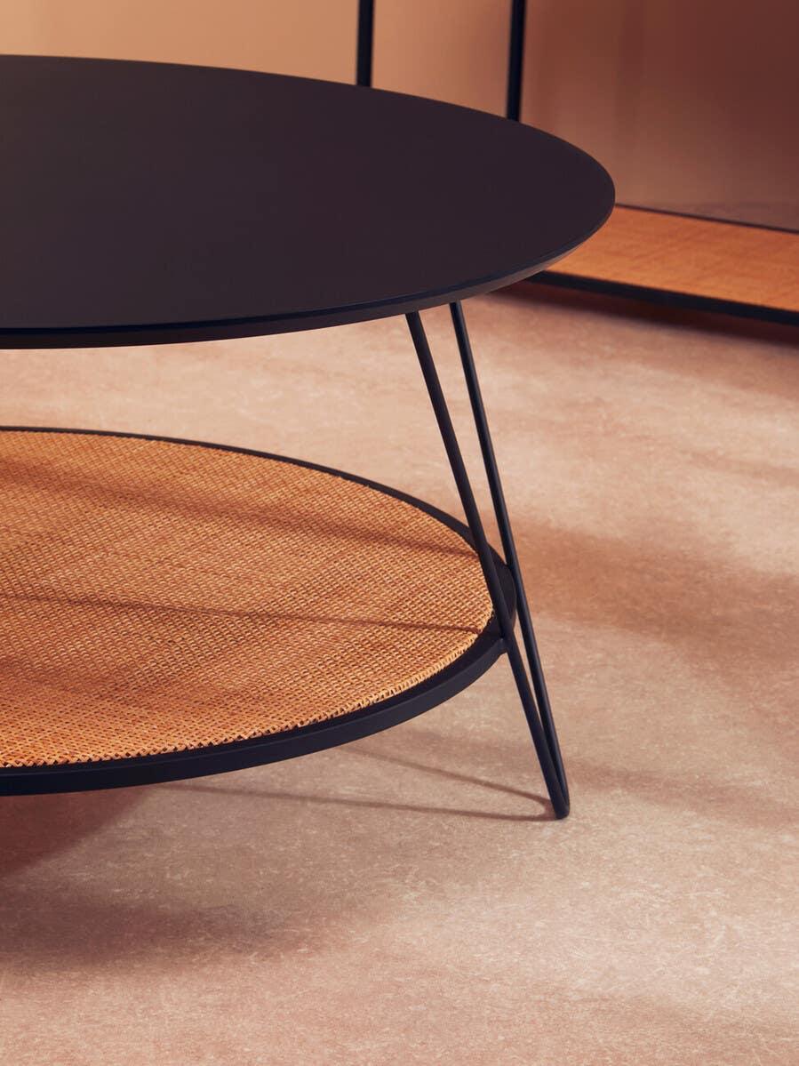 Round black coffee table with wire legs and rattan lower shelf on beige flooring
