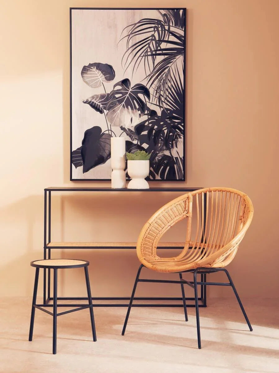 Rattan accent chair with black metal legs beside console table and monochrome leaf wall art