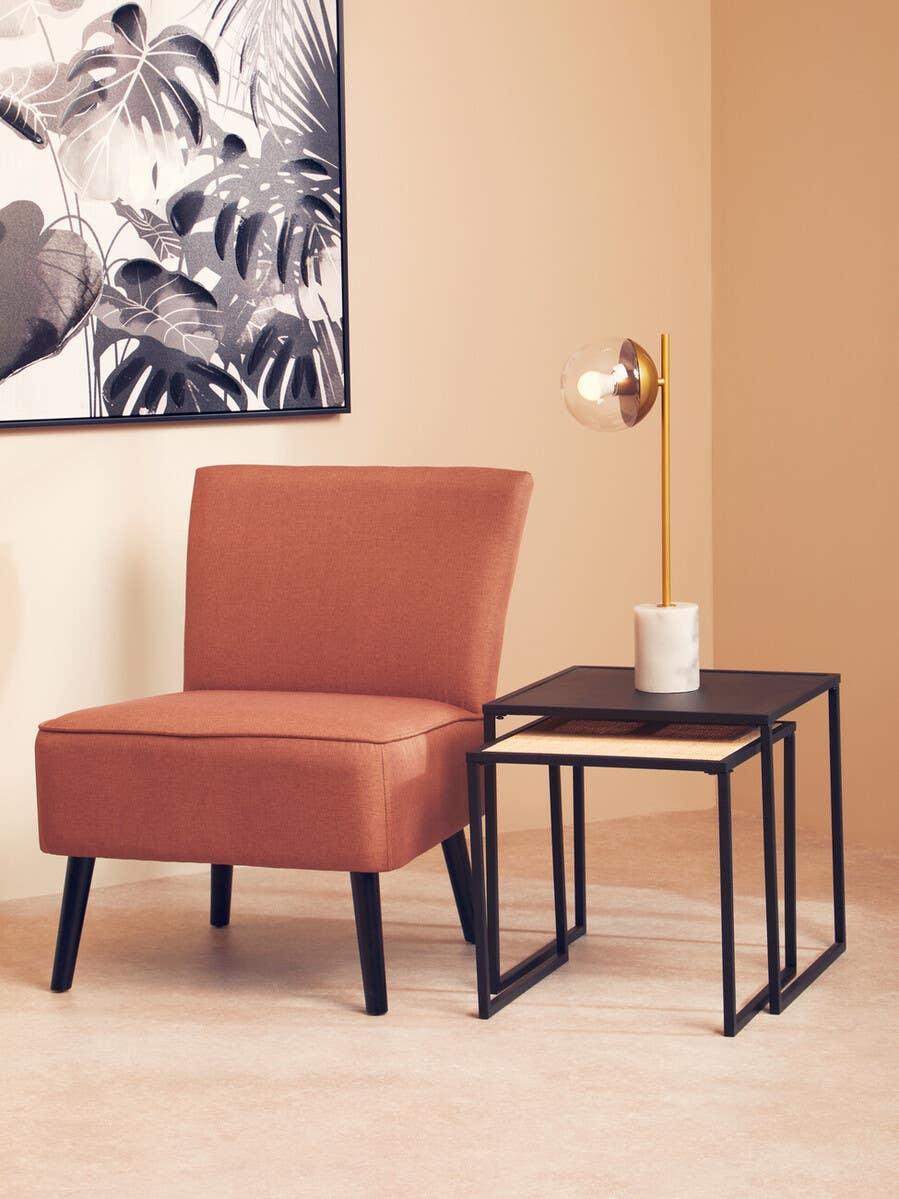 Modern accent chair with rust fabric, black legs beside nesting tables and gold table lamp