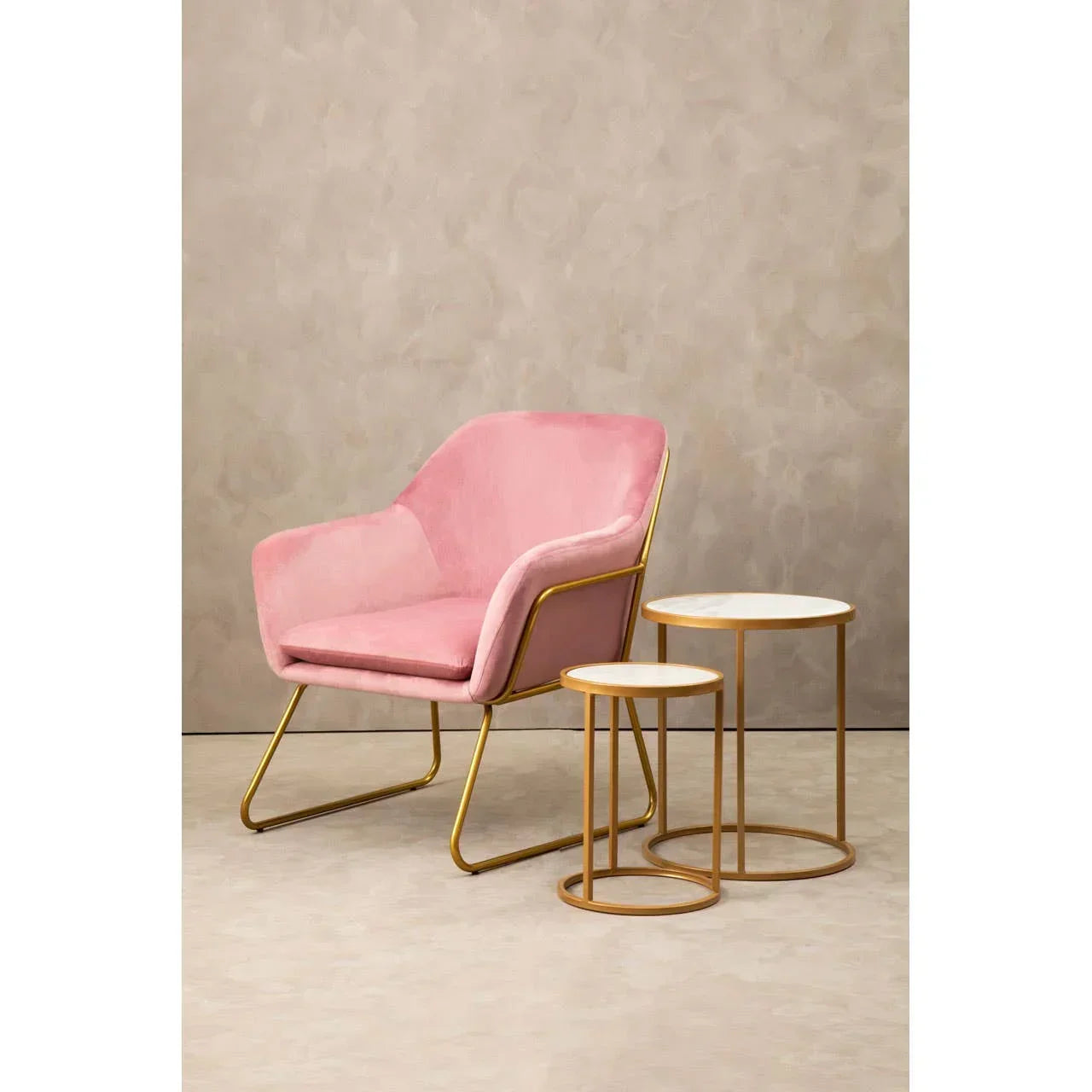 Pink velvet accent chair with gold legs beside two round marble top side tables