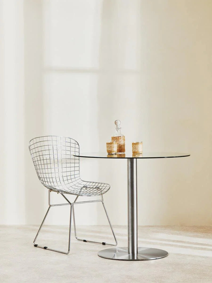 Glass dining table with metal pedestal base and wire chair in minimalist setting