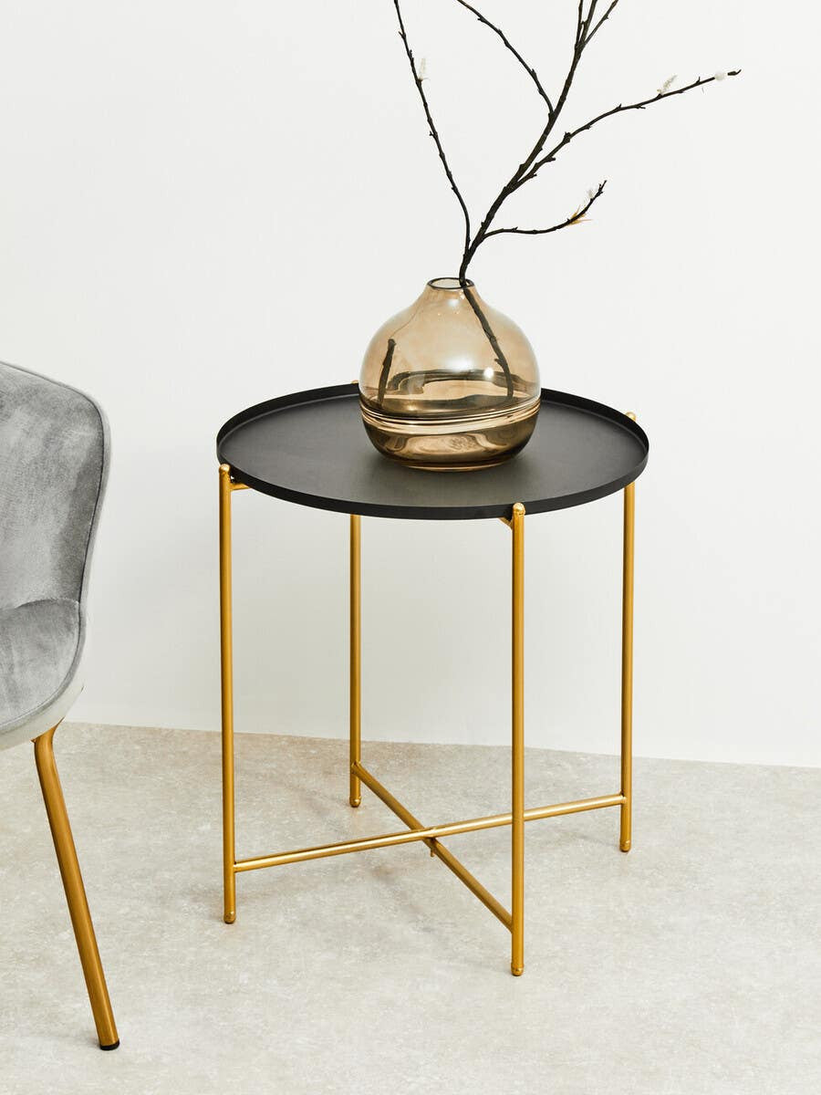 Round black top side table with gold metal legs, featuring a decorative vase with branches on top, positioned next to a gray upholstered chair on a light-colored floor