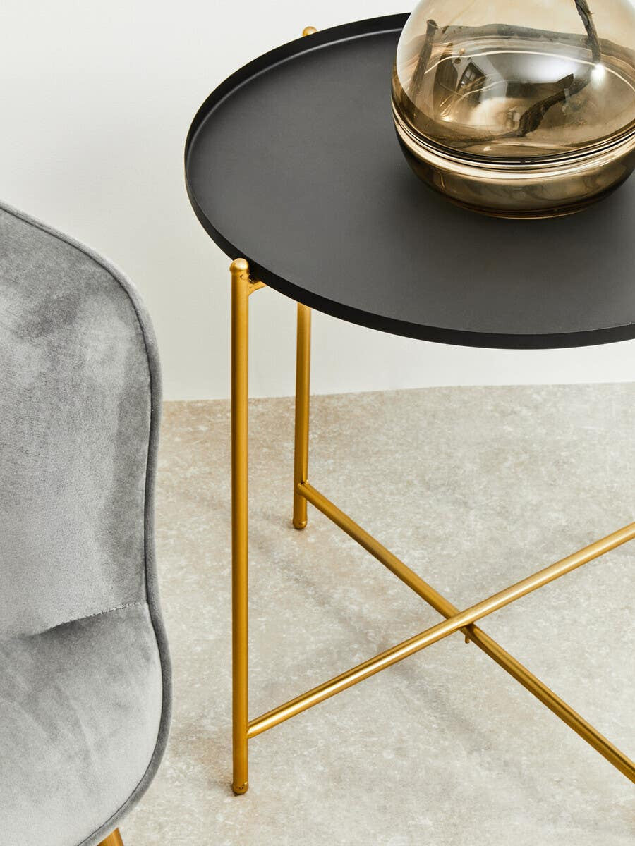 Black top side table with a round surface and gold metal legs, positioned next to a gray upholstered chair on a light-colored floor. A decorative glass vase sits on the table's surface