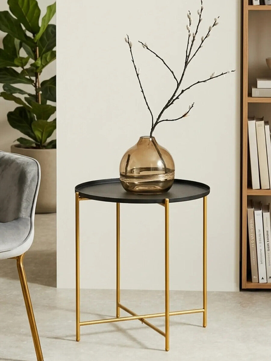 A black top side table with a round surface and gold metal legs, featuring a decorative vase with branches on top, set against a light-colored wall and a bookshelf in the background. A gray upholstered chair is partially visible to the left
