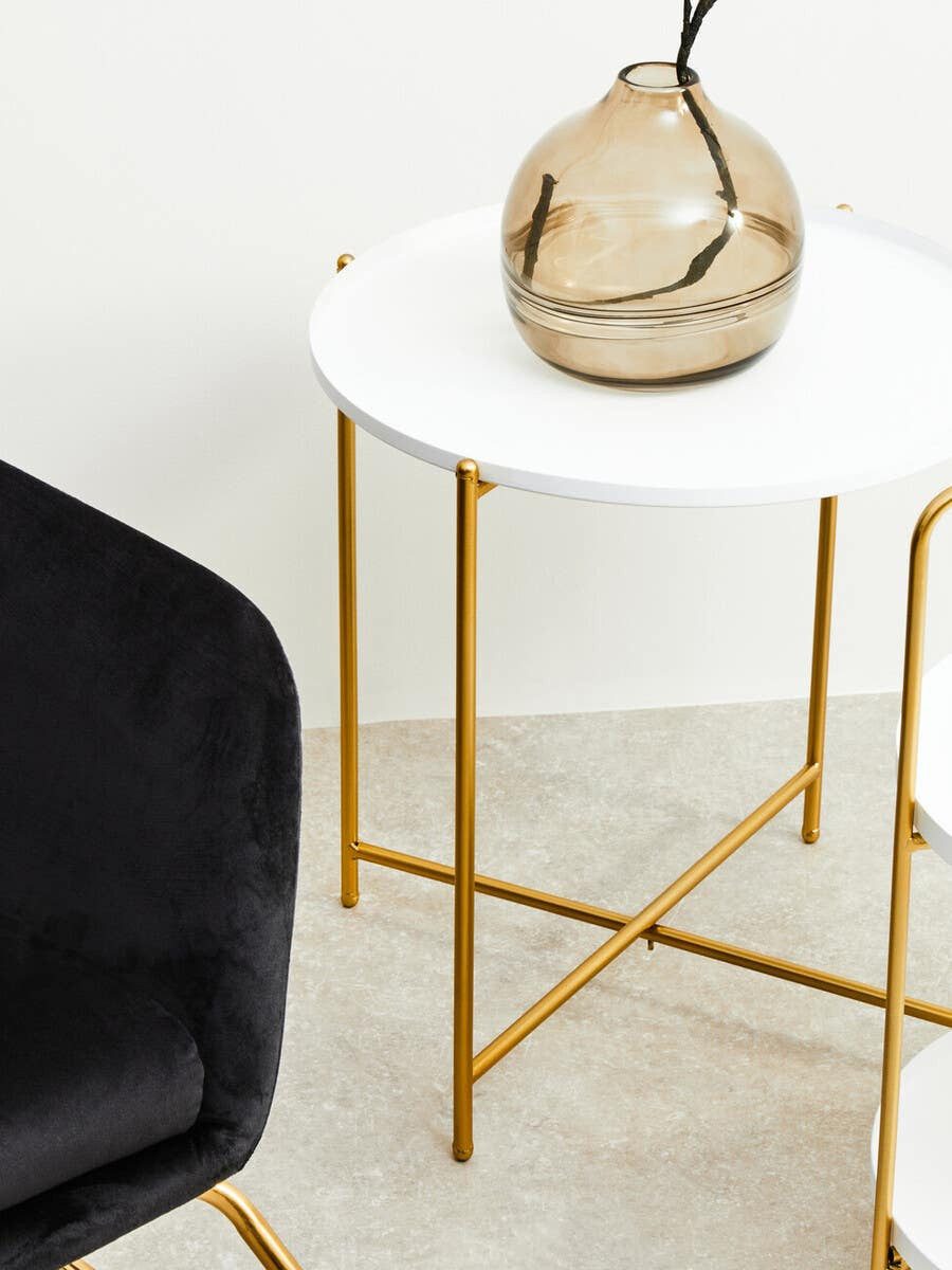 A white round side table with a smooth surface and gold metal legs, accompanied by a decorative glass vase on top. A black upholstered chair is partially visible beside the table, set against a light-colored floor
