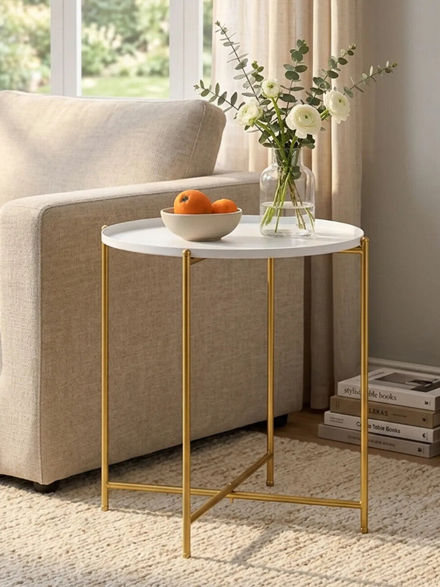 A white round side table with a smooth marble-like top and sleek gold metal legs, positioned next to a beige sofa. A bowl of oranges sits on the table, accompanied by a small vase with flowers. The setting features natural light coming through a nearby window, with a soft rug underneath
