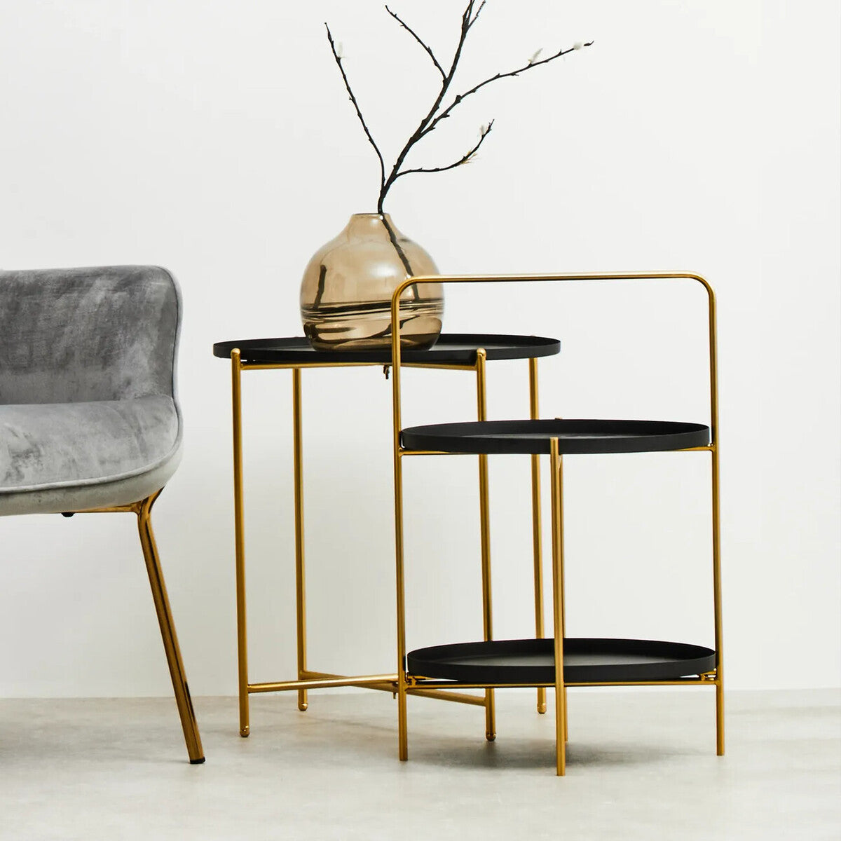 A two-tier side table featuring a sleek black top and a gold metal frame, showcasing a modern design. The table has two circular surfaces, with the upper tier slightly smaller than the lower one. A decorative vase with branches is placed on the top tier, while the table stands next to a gray upholstered chair with wooden legs. The background is a plain white wall, enhancing the table's elegant appearance