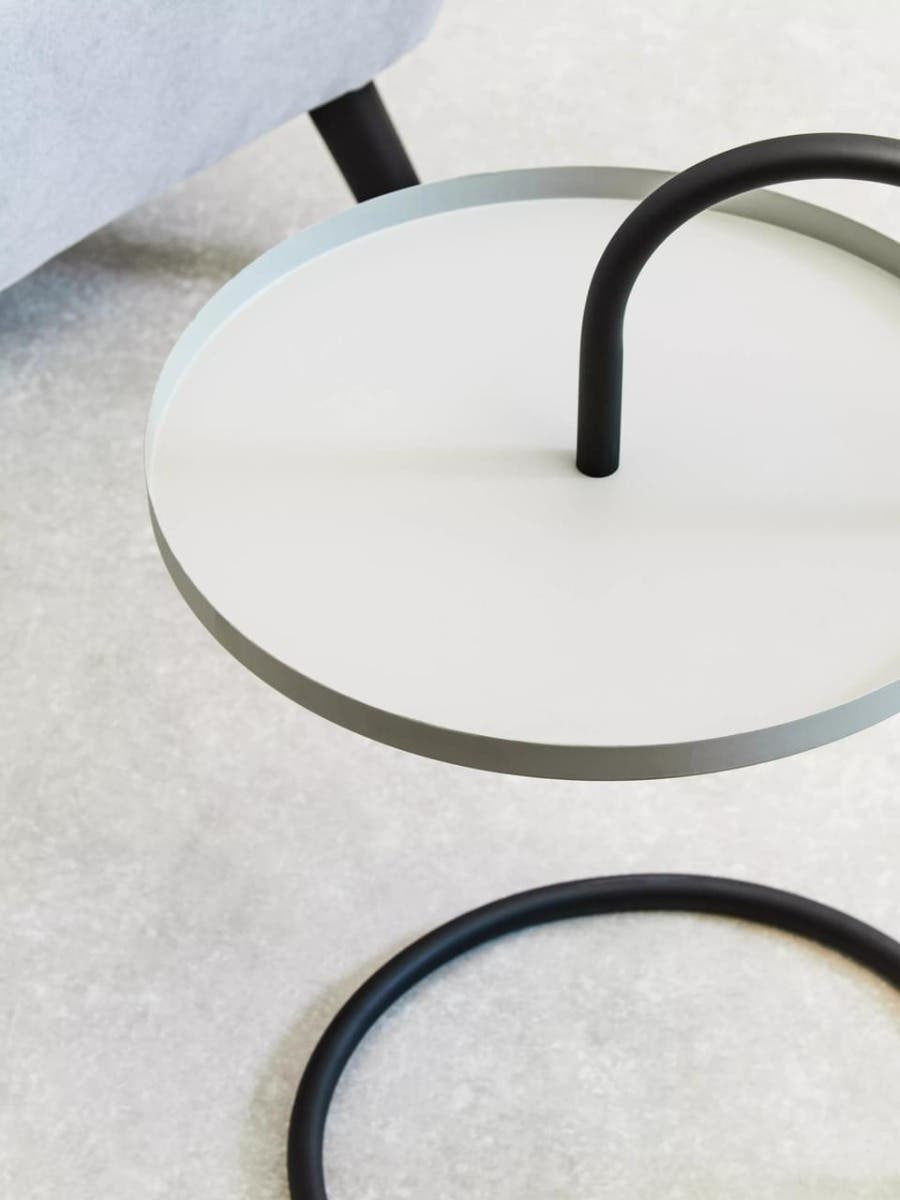 A round, grey and black side table featuring a sleek, minimalist design. The tabletop is circular and grey, supported by a black, curved metal frame that creates a hanging effect. The surface is smooth and modern, set against a light-colored background