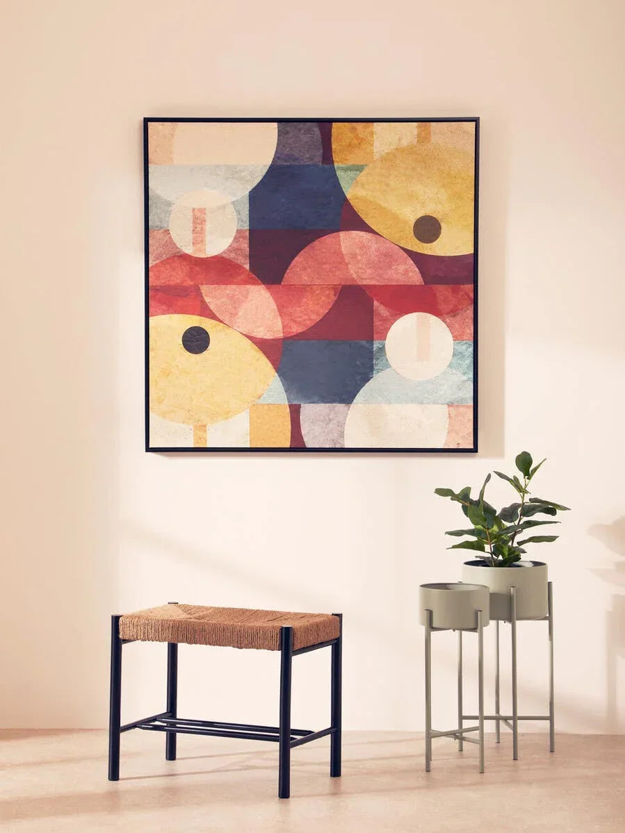Woven seat bench with black frame beside green plant stands and abstract wall art