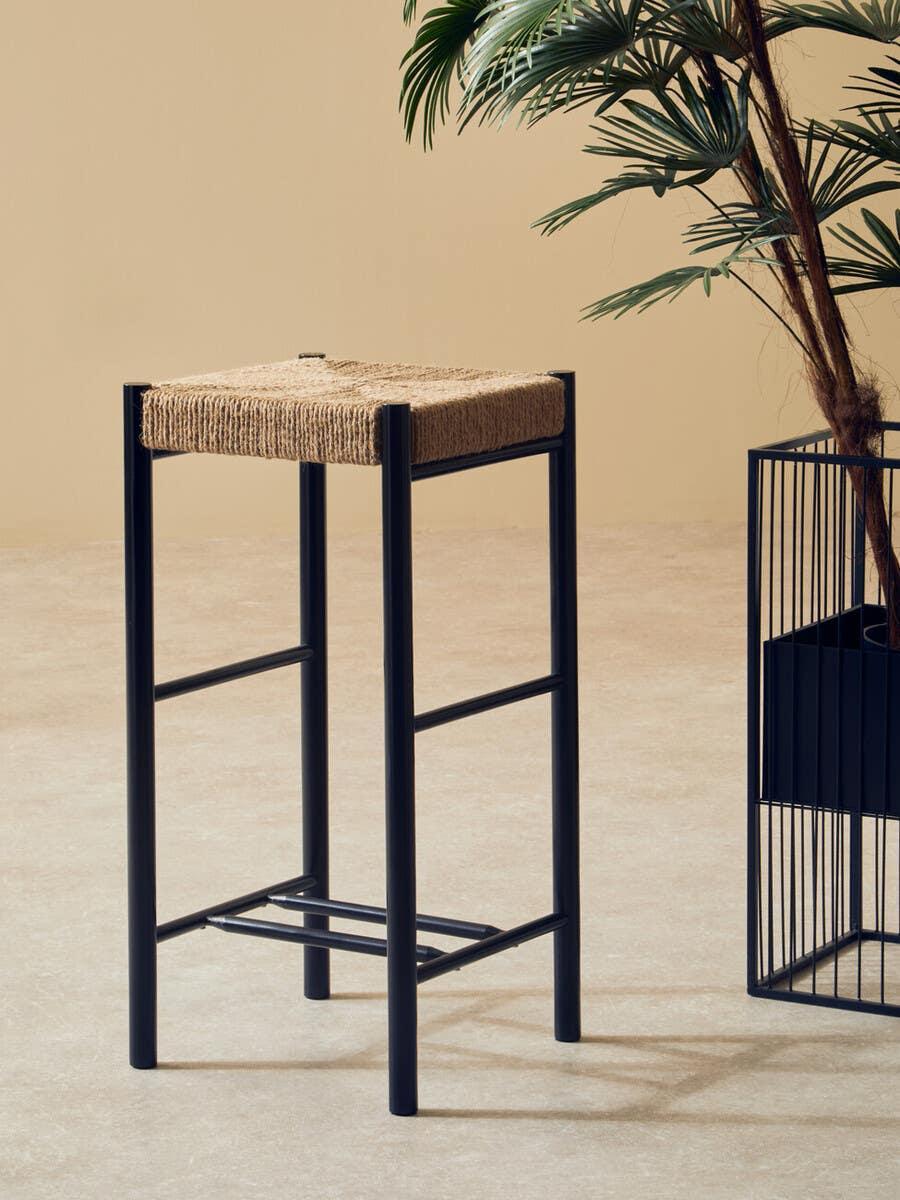 Black metal bar stool with woven natural seat beside indoor plant