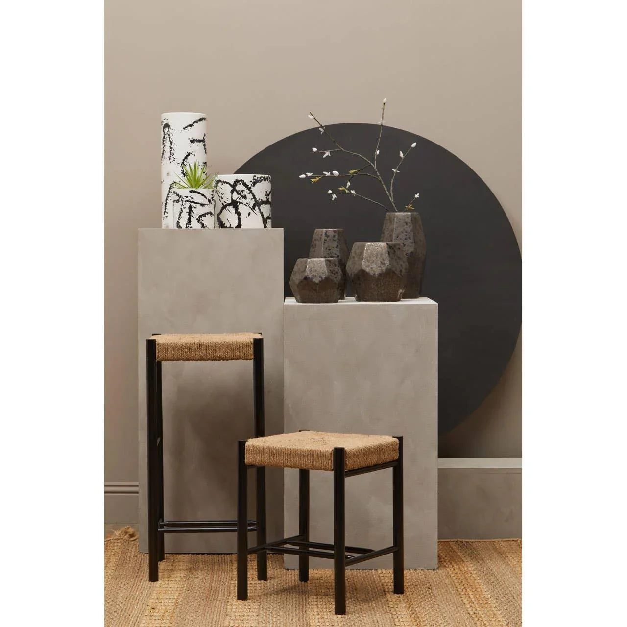 Set of two woven seat stools with black metal frames on natural jute rug, modern display vases