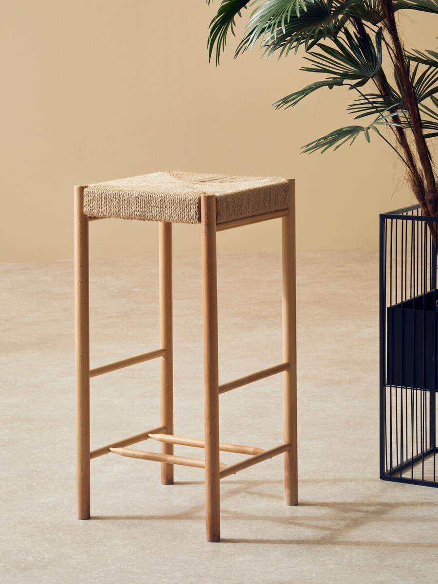 Natural wood bar stool with woven seat beside indoor plant