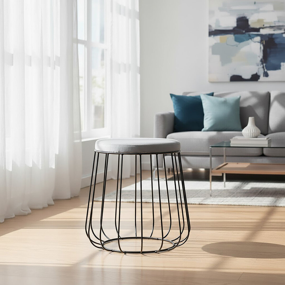 Grey velvet stool with a sleek black metal frame, positioned in a bright room with large windows. The stool features a soft, plush top and a modern, open design. In the background, a comfortable grey sofa with colorful throw pillows and a wooden coffee table are visible, along with light-colored flooring