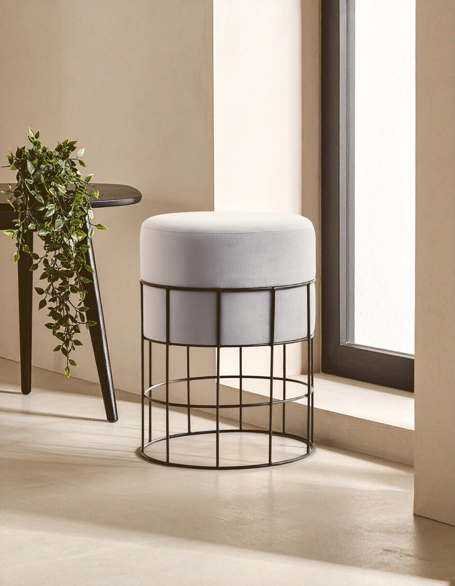 Velvet stool with a round top in light gray, supported by a black metal cage frame, positioned near a window with natural light illuminating the scene. A small plant is visible on a nearby table