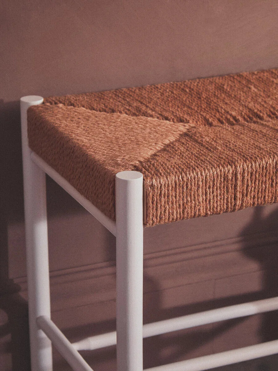 Woven seat bar stool with white metal legs against a muted brown wall