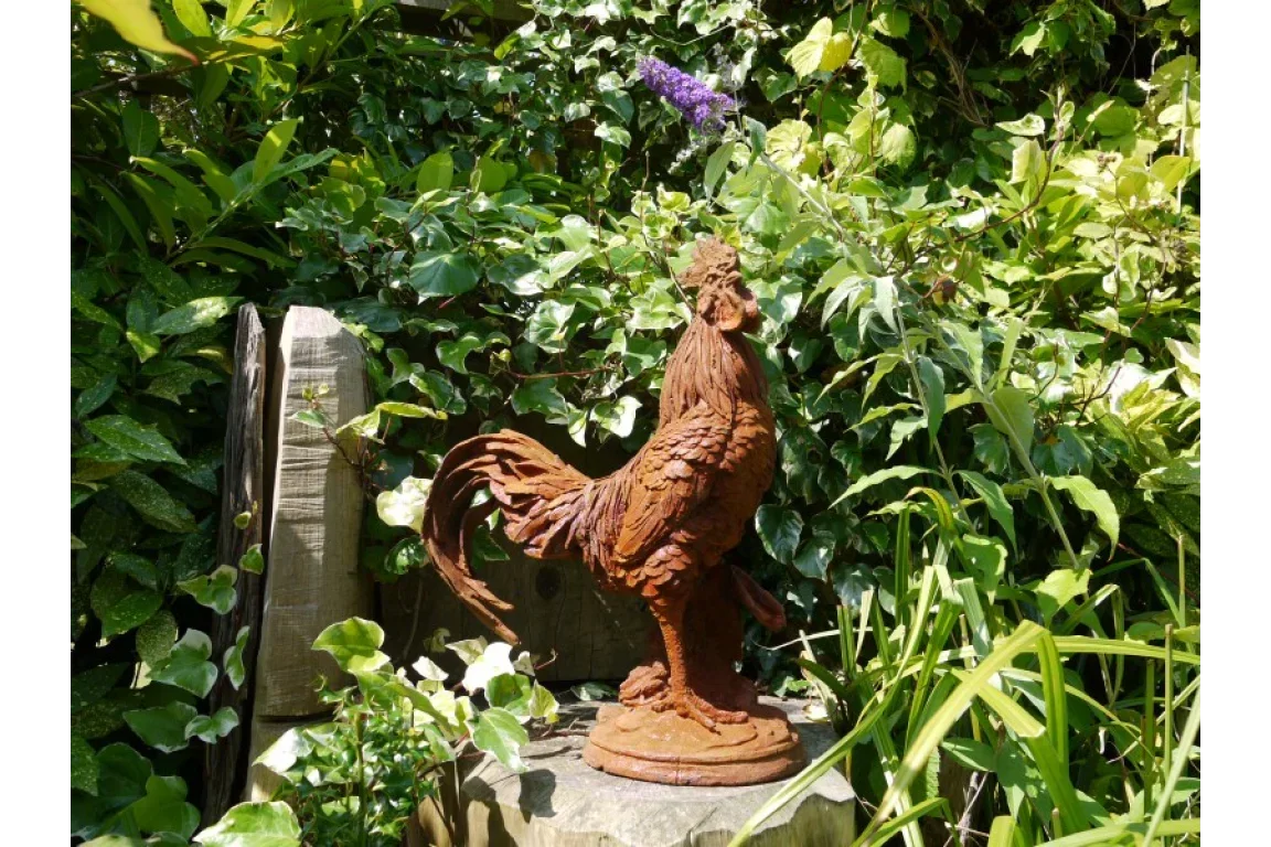 Rust effect rooster garden ornament on stone surrounded by lush green plants