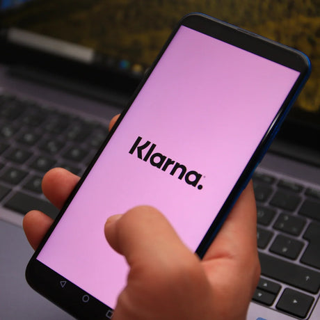 Hand holding smartphone displaying Klarna app screen in front of a laptop keyboard