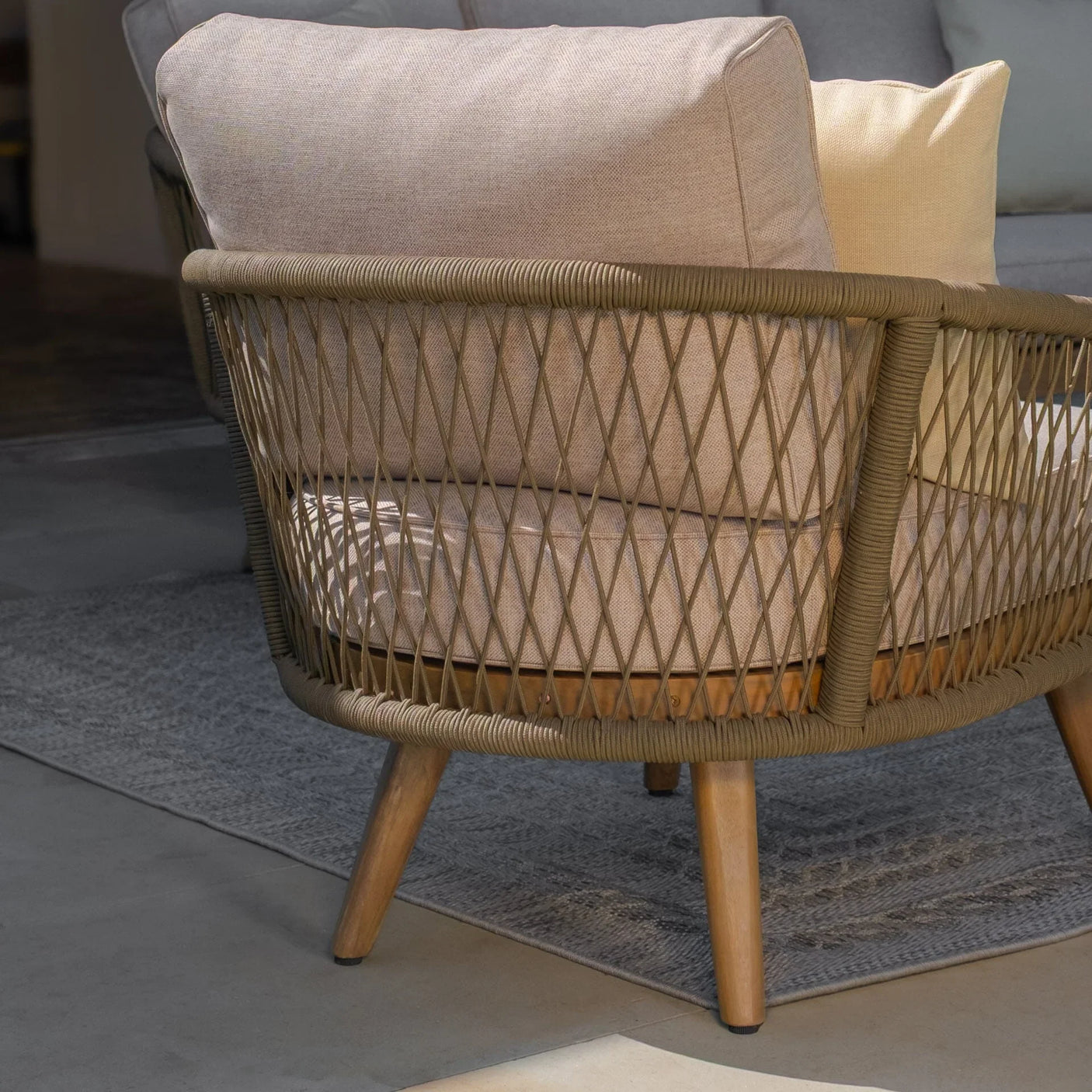Woven rattan armchair with beige cushions and wooden legs on a grey rug