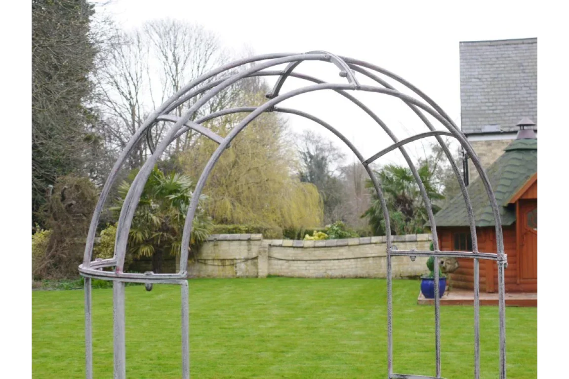Metal garden arch frame on lawn with stone wall and trees in background