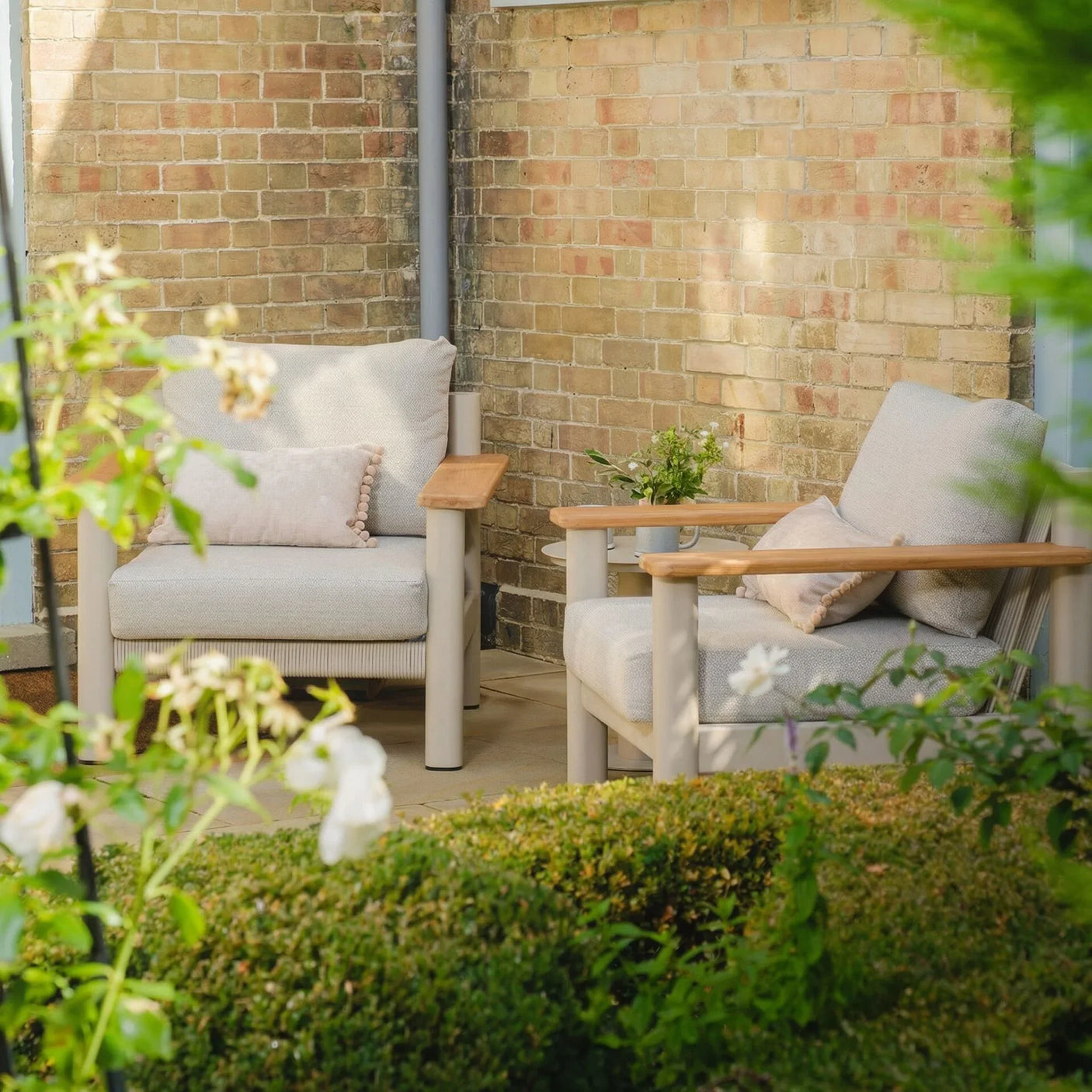 Outdoor garden lounge chairs with beige cushions and wooden arms by brick wall