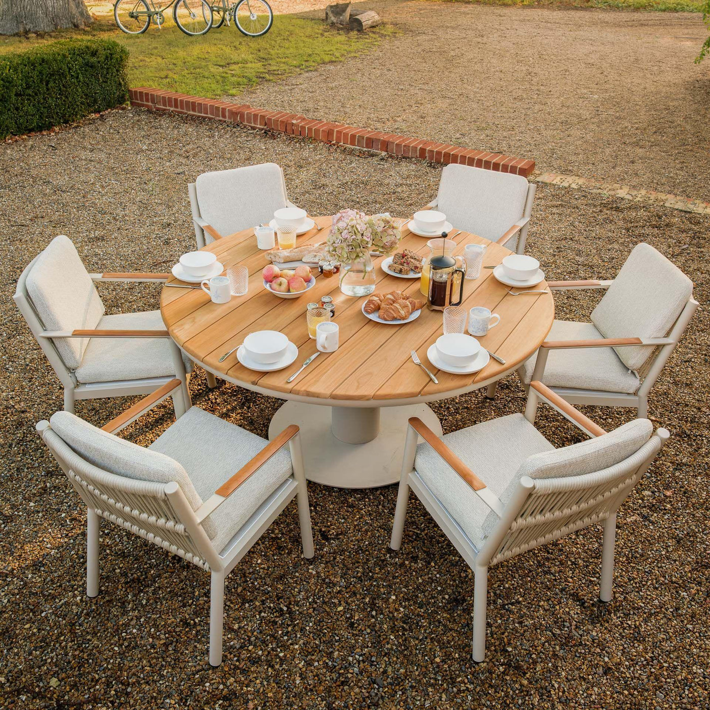 Round wooden outdoor dining table set with six cushioned chairs arranged for breakfast