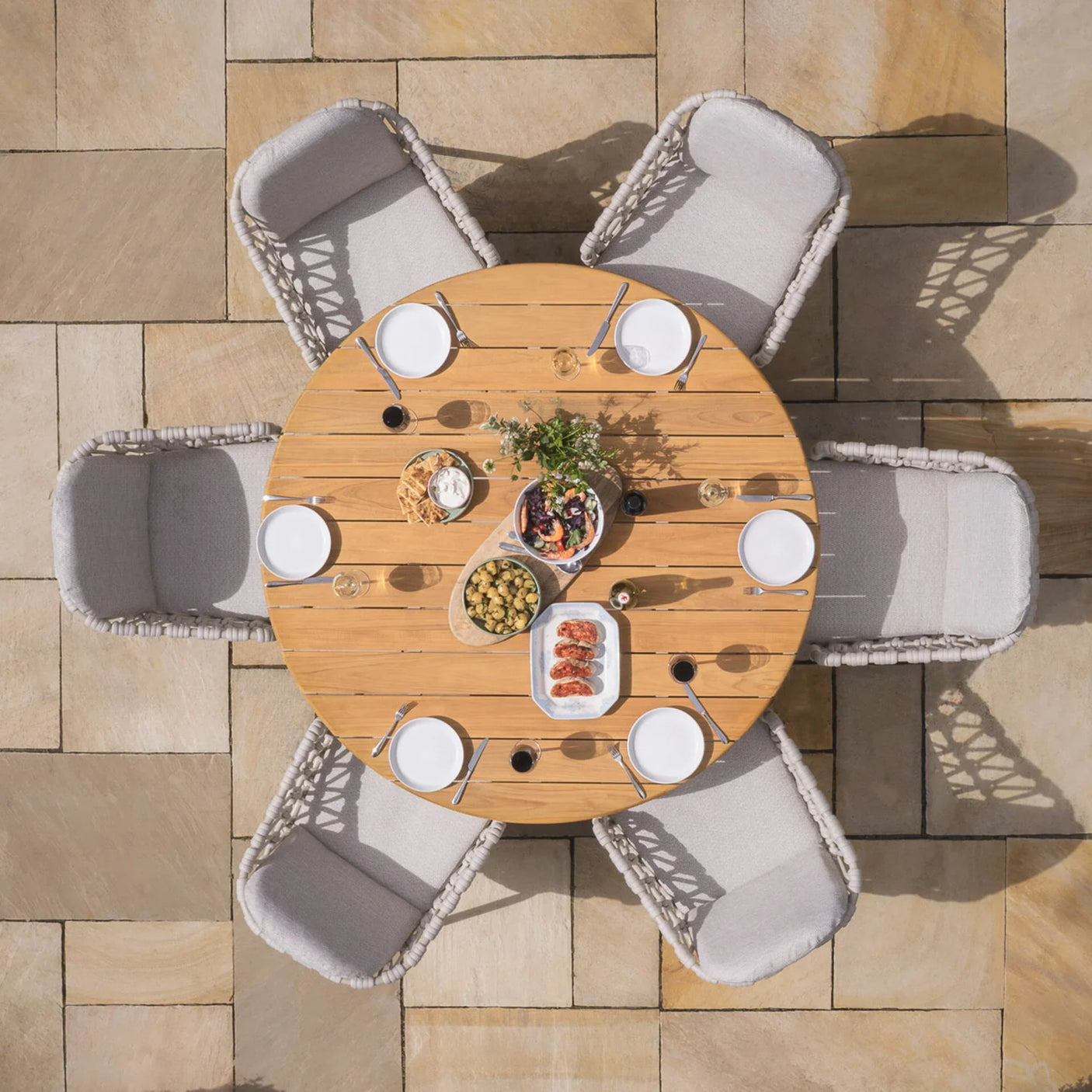 Round wooden outdoor dining table with six cushioned chairs set for a meal on stone patio