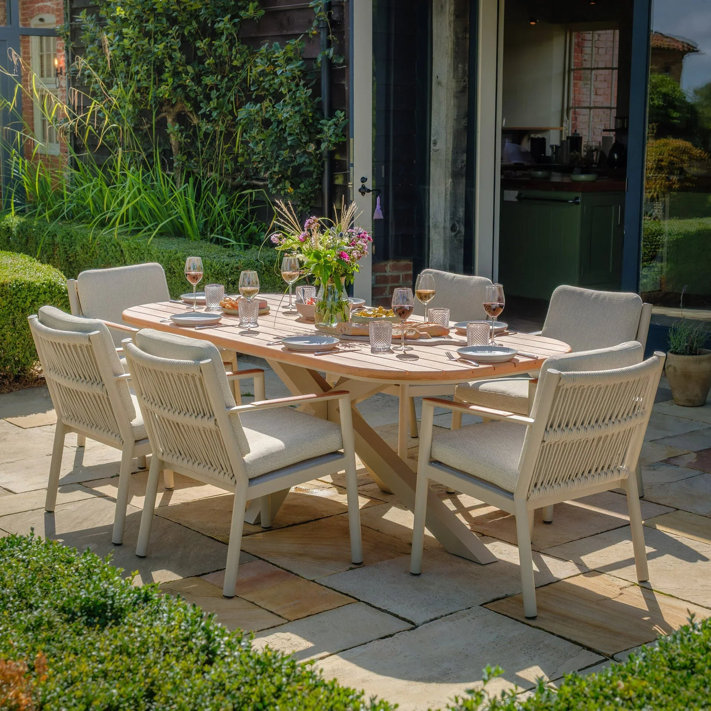 Outdoor wooden dining table set with six cream rope chairs on a stone patio, tableware arranged